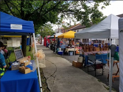 A vibrant outdoor market scene with colorful tents, vendors, and shoppers along a tree-lined pathway.