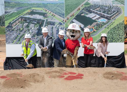 Groundbreaking ceremony with five people and a mascot, holding shovels in front of a construction site and project rendering.