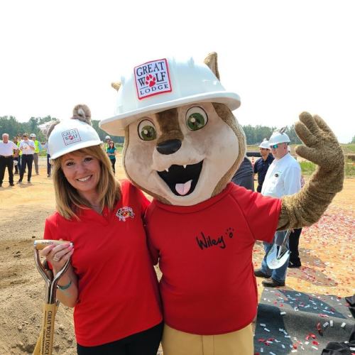 A woman and a wolf mascot in hard hats pose together at a groundbreaking event for Great Wolf Lodge.