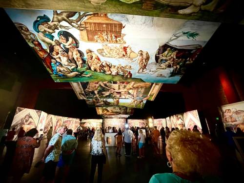 Visitors admire large, vibrant ceiling murals in an art exhibition, surrounded by illuminated wall displays.