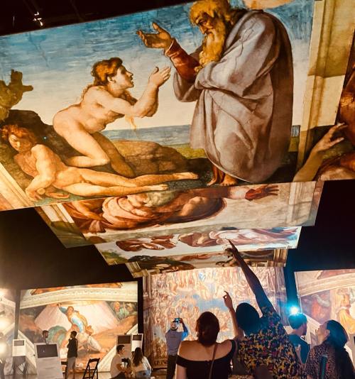 Visitors admire large-scale reproductions of Michelangelo's artwork displayed on the ceiling and walls of an exhibition.