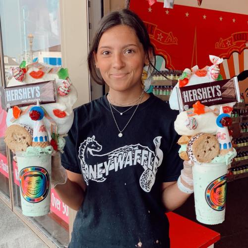 A young woman holds two oversized ice cream cups topped with candy and cookies, smiling in front of a colorful shop.