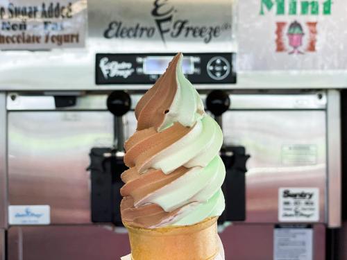 A swirl of chocolate and mint soft serve ice cream in a cone, with ice cream machines in the background.