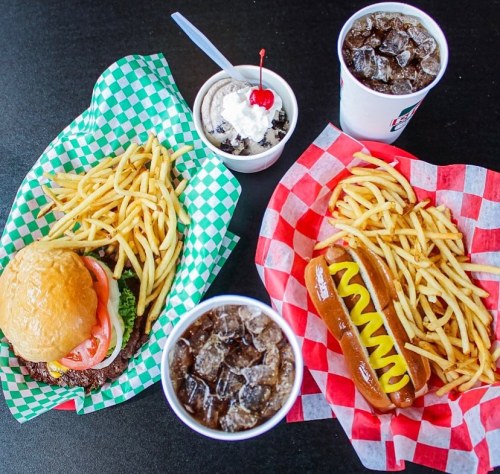 A burger with fries, a hot dog with fries, and two cups of soda, one topped with a cherry and whipped cream.