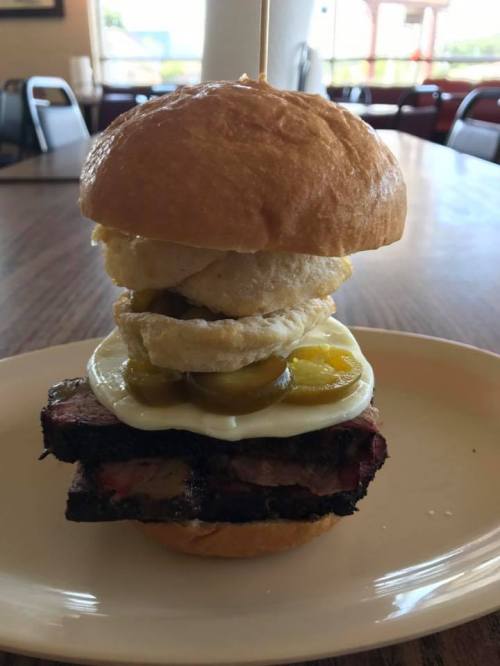 A stacked sandwich with brisket, pickles, fried egg, and a soft bun, served on a plate in a casual dining setting.