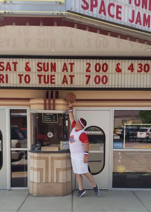 A person in a basketball jersey poses playfully with a basketball at a movie theater marquee displaying showtimes.