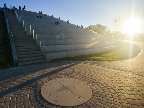 A sunlit park with stone steps and a compass rose on the ground, people sitting on the steps in the background.