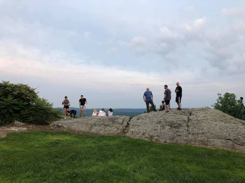 A group of people standing on a rocky outcrop with a grassy area, overlooking a scenic view under a cloudy sky.