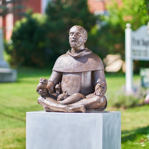 A bronze statue of a seated monk in meditation, surrounded by greenery and a sign in the background.