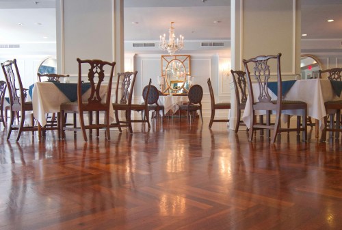 Elegant dining area with wooden floors, tables set with white linens, and a chandelier in the background.