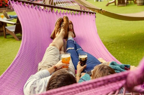 A couple relaxes in a pink hammock, enjoying drinks on a sunny day in a green outdoor setting.