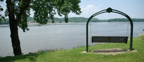A swing set by a river, surrounded by green grass and trees, under a clear blue sky.