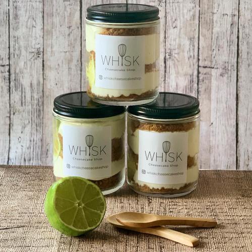 Three jars of cheesecake layered with graham cracker crust, topped with a lime, and wooden spoons beside them.