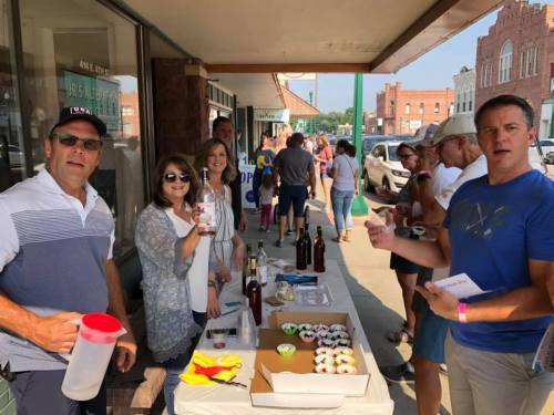 A group of people stands outside a shop, enjoying food and drinks during a community event on a sunny day.