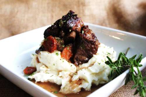 A plate of creamy mashed potatoes topped with tender beef and garnished with herbs and vegetables.