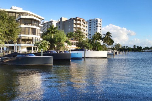 A serene waterfront scene with boats docked near modern buildings and palm trees under a clear blue sky.