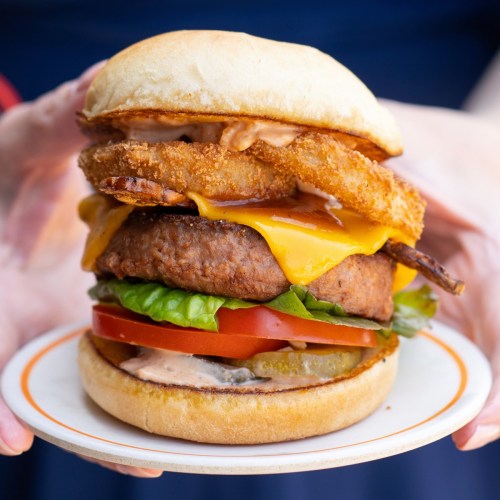 A close-up of a stacked burger with cheese, lettuce, tomato, pickles, and crispy onion rings on a plate.