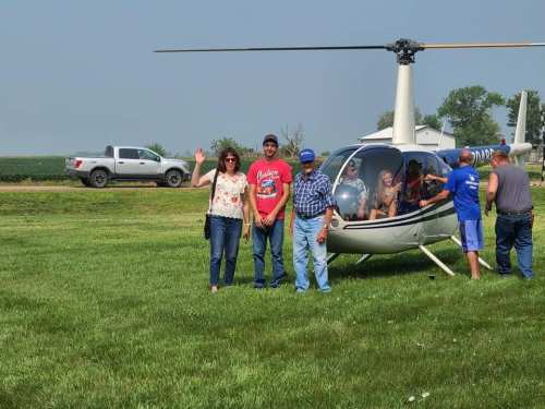 A group of people stands near a helicopter on a grassy field, with some waving and others seated inside.