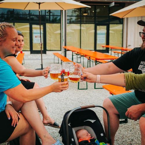 Four friends toast with beer mugs while seated at an outdoor table, with a baby in a stroller nearby.