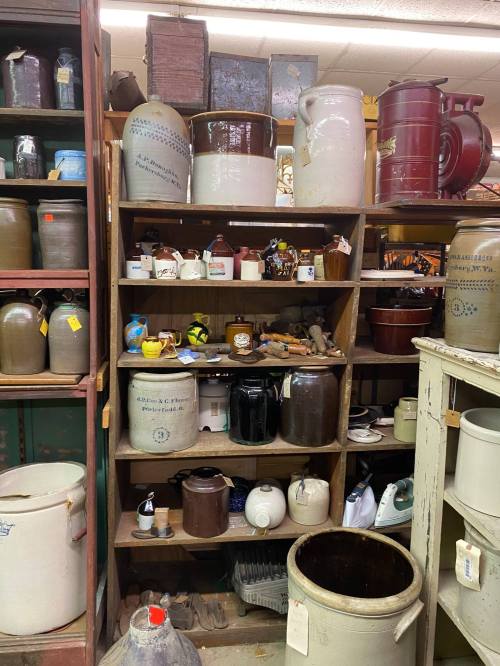 A wooden shelf filled with various antique jars, crocks, and pottery items in a vintage store.