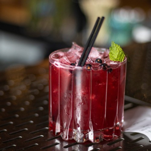 A refreshing red cocktail with ice, garnished with mint, served in a textured glass on a bar counter.