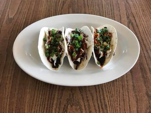 Three tacos on a white plate, filled with shredded meat, topped with green onions, on a wooden table.