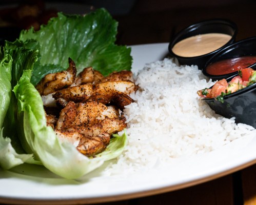 A plate featuring grilled shrimp wrapped in lettuce, served with rice, salsa, and a dipping sauce.