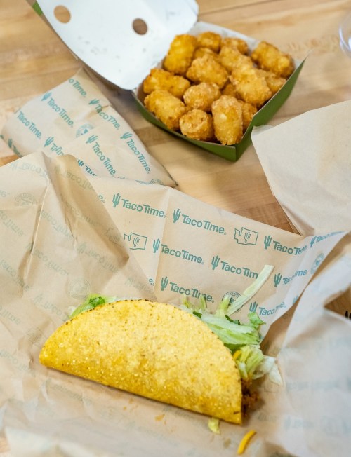 A taco with lettuce on a paper wrapper next to a box of golden tater tots on a wooden table.