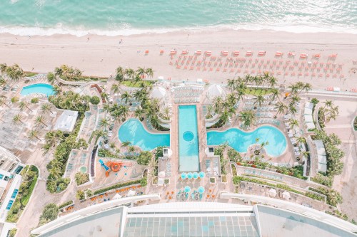 Aerial view of a resort with multiple pools, palm trees, and a beach with lounge chairs along the shoreline.