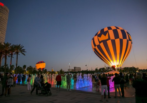 Colorful hot air balloons illuminate the night sky, surrounded by vibrant fountains and a crowd enjoying the scene.