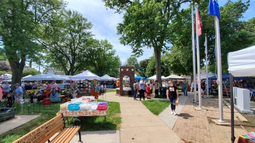 A bustling outdoor market with tents, vendors, and people walking along a path surrounded by trees.