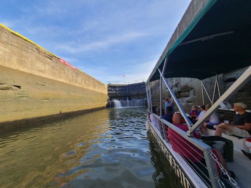 A boat approaches a lock with water cascading down, surrounded by stone walls and a clear blue sky.
