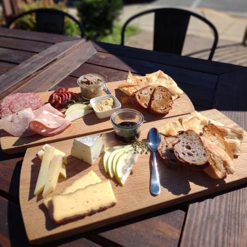 A wooden platter with assorted cheeses, cured meats, crackers, and fruit, set on a table outdoors.