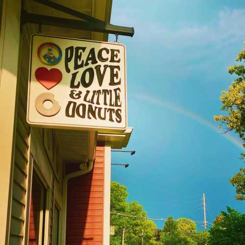 Sign for a shop reading "Peace Love & Little Donuts" with a rainbow in the background and a clear blue sky.