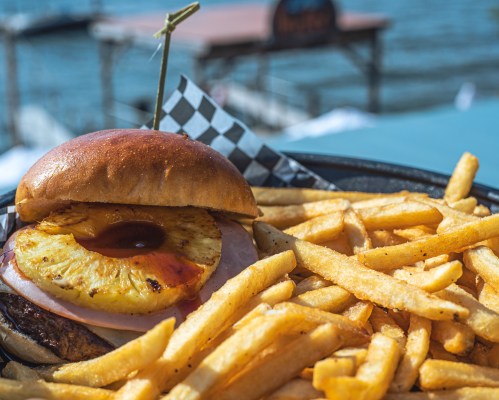 A close-up of a burger with a grilled pineapple slice, served with a side of golden fries, against a waterfront backdrop.