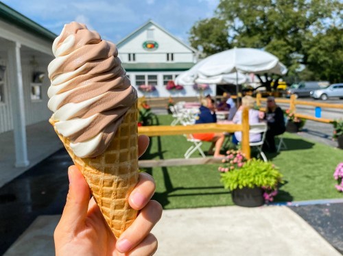 A hand holds a swirled chocolate and vanilla ice cream cone, with a café and outdoor seating in the background.