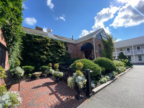A brick pathway leads to a building surrounded by lush greenery and flowers under a partly cloudy sky.