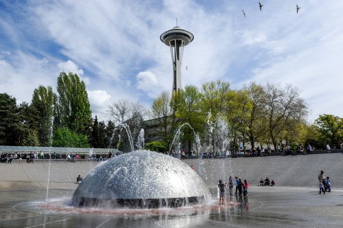 A fountain in a park with the Space Needle in the background, surrounded by trees and people enjoying the day.