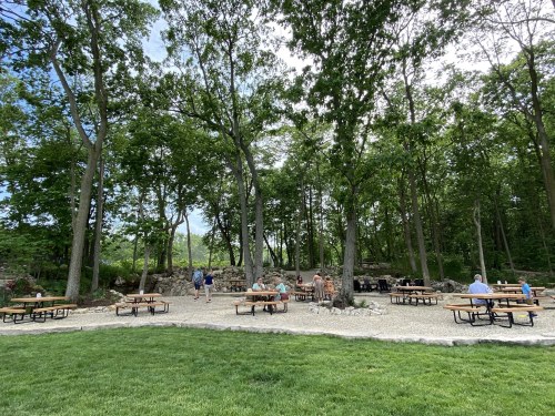 A scenic outdoor area with picnic tables surrounded by trees and people enjoying the space.
