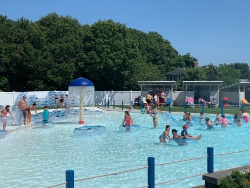 A busy water park scene with people enjoying a pool, floating on tubes, and a water feature under a clear blue sky.