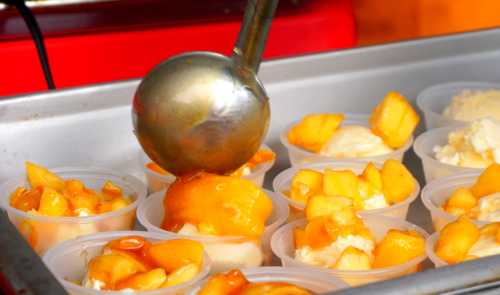 A metal scoop serving peach dessert into plastic cups filled with fruit and cream.