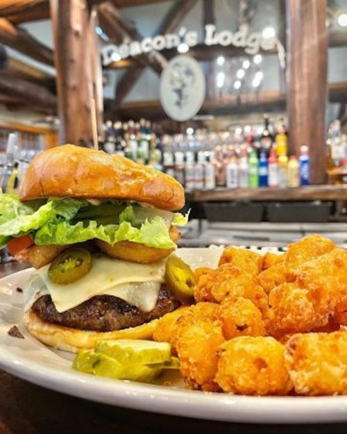 A juicy burger with lettuce, tomato, jalapeños, and cheese, served with crispy tater tots in a rustic bar setting.