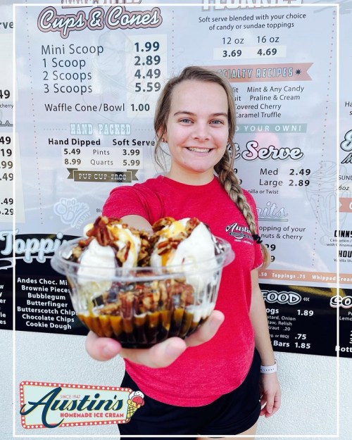 A smiling young woman holds a dessert cup with ice cream and toppings in front of an ice cream menu.