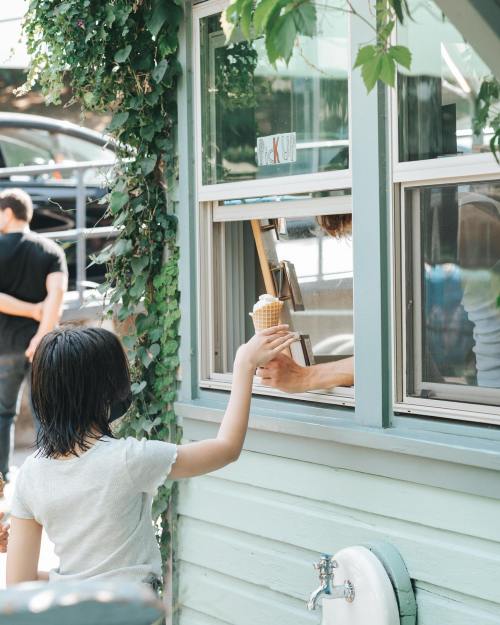 A child reaching for an ice cream cone from a window, surrounded by greenery and a bustling outdoor scene.