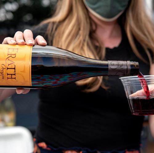 A person pours red wine from a bottle into a clear cup while wearing a mask.