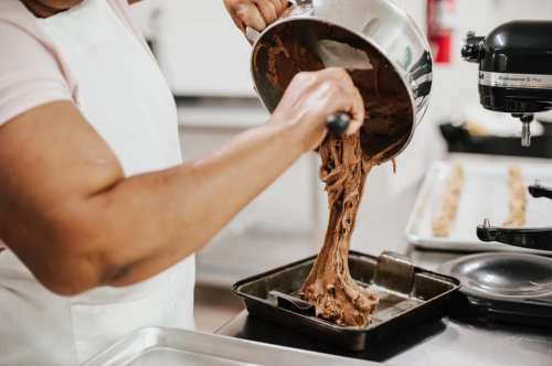 A person pours thick chocolate batter from a mixing bowl into a baking tray in a kitchen setting.