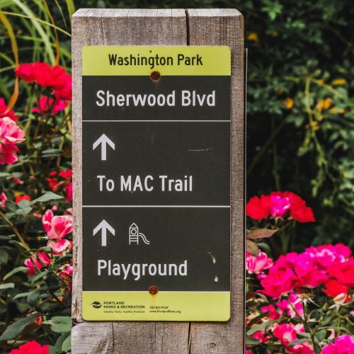 Signpost in Washington Park indicating directions to Sherwood Blvd, MAC Trail, and Playground, surrounded by flowers.