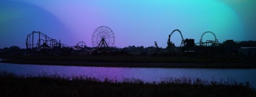 Silhouette of amusement park rides against a colorful twilight sky, reflected in a calm waterway.