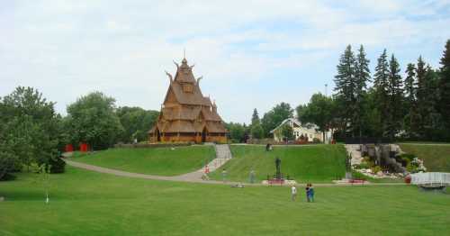 A wooden structure resembling a traditional Scandinavian church, surrounded by green grass and trees, with people walking nearby.