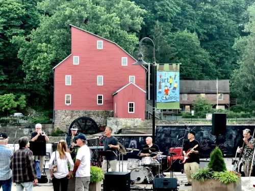 A band performs outdoors near a red barn, with trees and a banner in the background, as people enjoy the music.